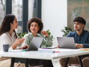 Corporate Teamwork. Three young colleagues of mixed ethnicity sitting at table with laptops in a modern office, collaborating on a project. Coworkers enjoying cooperative professional team work